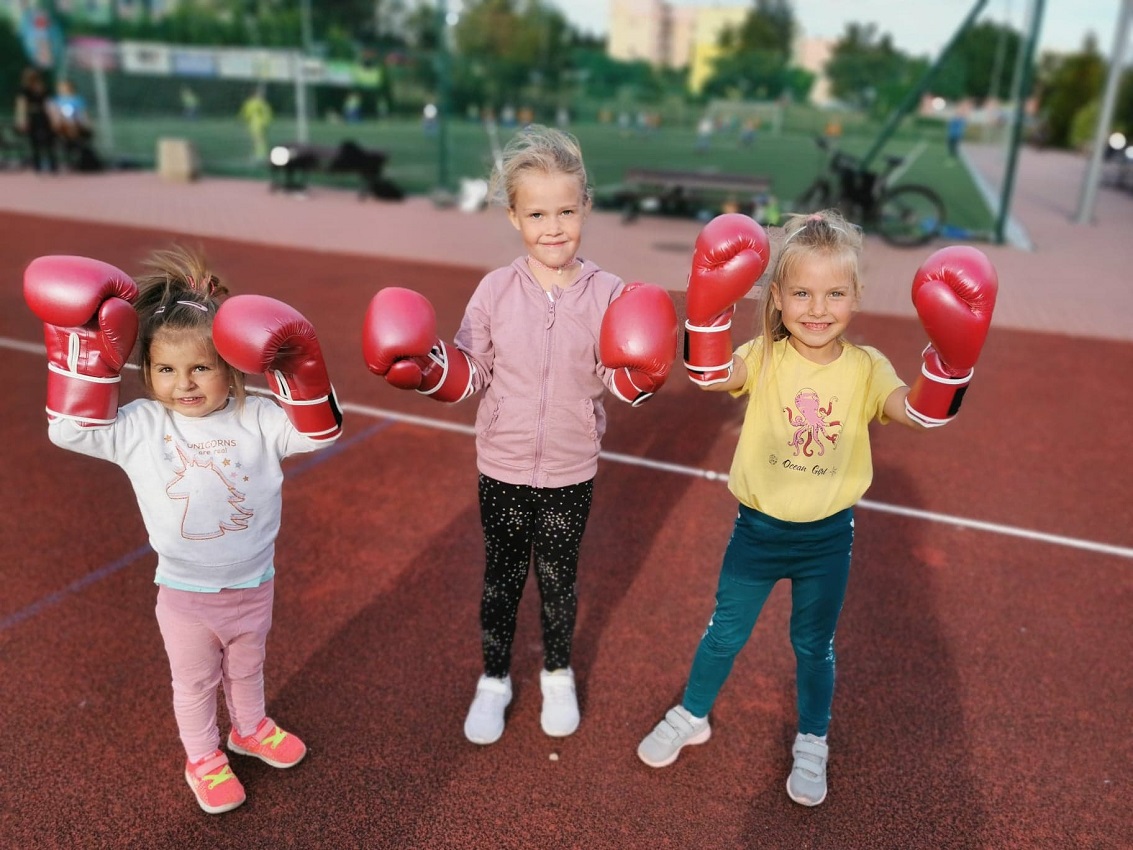 trzy dziewczynki stoja z uniesionymoi rękoma na płycie boiska piłkarskiego, mają ubrane rękawice bokserskie
